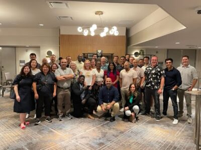 A group of people is gathered in a well-lit room, posing for a group photo. They are standing and kneeling, smiling at the camera. The room has modern decor with a light fixture on the ceiling and a wooden panel in the background.